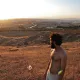 L'image montre un homme debout sur une colline, regardant vers un paysage vallonné au coucher du soleil. Il a les cheveux et la barbe bruns, et porte un t-shirt. Le terrain autour de lui est sec et couvert d'herbes hautes. En arrière-plan, on peut voir un petit village et des champs qui s'étendent à perte de vue, créant un panorama paisible et naturel. La lumière du soleil met en valeur les couleurs dorées de la scène.