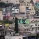 L'image montre un paysage urbain d'une ville, où plusieurs bâtiments sont alignés sur une colline. À travers ces constructions, on peut voir de grandes peintures murales représentant des yeux, aux couleurs vives, qui ajoutent une dimension artistique à cet environnement. Les bâtiments varient en couleurs et en textures, allant du beige au rose, et certains semblent en cours de rénovation. L'atmosphère générale évoque un mélange de quotidien et de créativité, avec une forte présence d'art urbain.