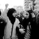 L'image montre un groupe de personnes lors d'une manifestation. Deux jeunes femmes, l'une portant un hijab et l'autre les cheveux détachés, semblent échanger des mots tout en levant les bras, ce qui indique un sentiment d'engagement et de solidarité. L'ambiance est dynamique, et le noir et blanc donne une atmosphère intense à la scène. En arrière-plan, on peut apercevoir d'autres manifestants, créant une impression de mouvement et de rassemblement.