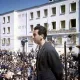 L'image montre un homme se tenant derrière un microphone, semblant s'adresser à une grande foule rassemblée devant lui. L'arrière-plan présente un bâtiment de style architectural simple avec plusieurs fenêtres, possiblement dans un contexte historique ou politique. Les membres de la foule semblent attentifs à son discours. L'éclairage naturel donne une ambiance claire à la scène, suggérant qu'elle pourrait avoir été prise en plein jour.