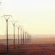 L'image montre une perspective sur un paysage de campagne, dominé par des pylônes électriques s'étendant sur une longue distance. Le sol est principalement sec et herbeux, avec des teintes dorées, suggérant la lumière douce d'un lever ou d'un coucher de soleil. En arrière-plan, on peut apercevoir des collines ou des montagnes floues, contribuant à une ambiance calme et désertique. L'ensemble évoque un sentiment d'immensité et de tranquillité.