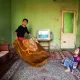 L'image montre une pièce modeste avec des murs verts. Au centre, deux enfants sont présents. L'un d'eux, un garçon, tient une grande couverture qu'il semble s'apprêter à lancer ou à étendre. L'autre enfant, une petite fille, est assise sur le sol, regardant le garçon avec curiosité. On peut voir un téléviseur allumé sur une petite table, et quelques vêtements accrochés au mur. Le décor est simple, avec un tapis et des canapés. L'ambiance générale semble familiale et décontractée.