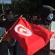 L'image montre un groupe de personnes participant à une manifestation. Au premier plan, un homme tient un drapeau tunisien rouge avec un croissant et une étoile blanche. D'autres manifestants portent des masques, et l'un d'eux tient une pancarte sur laquelle est écrit un message en anglais, notamment « IDEAS CANNOT BE DESTROYED ». L'arrière-plan est constitué d'arbres et de rues, ce qui donne un air de protestation pacifique.