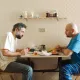 L'image montre deux hommes assis à une table dans une pièce au décor simple. Ils semblent en pleine conversation, chacun portant une expression sérieuse. Sur la table, il y a des assiettes, des verres et des aliments. L'un des hommes porte un t-shirt gris, tandis que l'autre est vêtu d'un t-shirt bleu. La pièce est éclairée de façon naturelle, et le mur est décoré d'un motif en damier. L'ambiance semble à la fois intime et chargée.