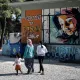 L'image montre un groupe de personnes marchant sur un chemin en gravier. À gauche, on peut voir un conteneur à ordures et à droite, un mur peint avec des graffitis colorés, dont le mot "Welcome to Ritsona" et le portrait d'une fille. Les personnes présentes semblent être une mère accompagnée de deux enfants, l'un d'eux tenant la main de la femme. L'environnement végétal suggère un cadre tranquille, malgré la situation.