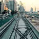 L'image montre une vue urbaine d'une ville moderne. On peut voir des routes animées avec des voitures circulant, ainsi qu'un important réseau de rails de train. À droite, il y a des bâtiments imposants et modernes qui reflètent l'architecture contemporaine. L'atmosphère est lumineuse, suggérant un moment de la journée proche du coucher du soleil avec des lumières douces. Les rails semblent bifurquer, suggérant une intersection, tandis que des travaux de construction ou d'aménagement sont visibles au premier plan.