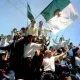 L'image montre une foule rassemblée, levant des bras et brandissant des drapeaux, dont celui de l'Algérie. Les personnes semblent joyeuses et engagées, célébrant un moment fort. L'atmosphère pourrait évoquer une célébration de l'indépendance ou un événement historique significatif pour le pays. Les visages sont animés et l'énergie collective est palpable.