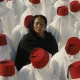 L'image montre un groupe de personnes vêtues de longues robes blanches, portant des fez rouges. Au centre, une femme en noir se tient les yeux fermés, contrastant avec l'uniformité des autres. Cette composition crée une atmosphère de méditation ou de réflexion, avec un fort accent sur la spiritualité et la communauté. Les couleurs et les vêtements évoquent une tradition culturelle riche.