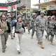 L'image montre un groupe de soldats en uniforme militaire marchant dans une rue. Ils semblent être dans une zone urbaine, possiblement dans un pays en conflit. Au premier plan, on voit une femme en vêtements civils, portant un gilet pare-balles, marchant à côté d'un homme également en tenue civile. Le décor autour d'eux comprend des bâtiments, des panneaux et d'autres signes de vie urbaine. L'atmosphère semble sérieuse, reflétant un environnement potentiellement tendu.