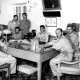 L'image montre un groupe de six hommes en uniforme militaire réunis autour d'une grande table en bois dans un bureau. Certains d'entre eux sont assis, tandis que d'autres se tiennent derrière. L'environnement décoré de manière classique inclut des éléments tels qu'une lampe de bureau et des tableaux accrochés au mur. L'atmosphère semble formelle, et les expressions des hommes varient entre concentration et sérieux.