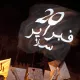 L'image montre une manifestation nocturne. À l'avant-plan, une personne tient un drapeau noir portant des inscriptions en blanc. Le drapeau semble être un symbole de protestation. En arrière-plan, on distingue des panneaux de revendications, également en arabe. L'atmosphère semble dynamique et engagée, illustrant un mouvement de contestation. La présence des palmiers suggère que l'événement se déroule dans un pays à climat chaud, probablement en Afrique du Nord.