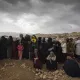 L'image montre un groupe de femmes rassemblées, probablement en attente de quelque chose. Elles portent des vêtements sombres, et certaines tiennent des enfants. Le ciel est nuageux, ce qui donne une ambiance sombre à la scène. En arrière-plan, on aperçoit des maisons sur une colline, soulignant un environnement rural ou de village. L'attente et la solidarité féminine semblent être des thèmes présents dans cette image.