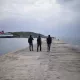 L'image montre trois personnes marchant sur un quai. Le ciel est nuageux, et l'atmosphère est calme. À l'arrière-plan, on peut voir un ferry amarré et une petite île. Le quai est en béton, et l'eau semble calme, reflétant les nuages. Les silhouettes des gens ajoutent une touche de vie à cette scène paisible.