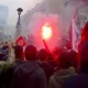 L'image montre une foule rassemblée dans une manifestation. Des personnes tiennent des drapeaux, dont un drapeau tunisien, et certains agitent des fumigènes qui dégagent de la fumée rouge. L'ambiance semble énergique et engagée, avec des bâtiments en arrière-plan. La foule paraît déterminée et unie autour de leur cause.