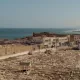 L'image montre un panorama d'une ville côtière, probablement située au bord de la mer Méditerranée. Au premier plan, on peut voir un cimetière avec de nombreuses tombes en pierre, disposées de manière régulière. À l'arrière-plan, il y a des bâtiments blancs, typiques des architectures méditerranéennes, qui bordent la côte. La mer s'étend à l'horizon, avec des vagues visibles, et il semble y avoir une légère brume sur l'eau. La scène dégage une ambiance paisible et sereine, avec une lumière douce évoquant l'heure du soir ou du matin.