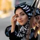 L'image montre une femme portant un hijab à motifs zebrés noirs et blancs. Elle se tient près d'une voiture, regardant vers la caméra avec une expression calme et confiante. Ses lunettes de soleil sont posées sur le haut de sa tête, ajoutant une touche de style. En arrière-plan, on peut apercevoir des contours flous d'un environnement urbain, suggérant un jour ensoleillé.