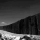 L'image montre un mur imposant s'étendant à l'horizon, dans un paysage désertique. Le mur est composé de grandes plaques de béton, parfois inégales. Au premier plan, il y a des débris et des éléments naturels, ce qui renforce l'impression d'un environnement aride et abandonné. Le ciel en arrière-plan semble dégagé, mais la scène est en noir et blanc, ce qui ajoute une atmosphère sombre et sérieuse à l'image.