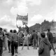 L'image montre une scène de rue animée, probablement lors d'une manifestation ou d'un rassemblement. Des groupes de personnes, en majorité des jeunes, se rassemblent autour d'une voiture. Certains tiennent des drapeaux ou des banderoles sur lesquelles des messages sont affichés, exprimant des revendications. L'atmosphère semble dynamique et chargée d'émotion, avec des visages montrant diverses réactions. En arrière-plan, on aperçoit un paysage urbain avec des arbres et des bâtiments. Le noir et blanc de la photo accentue le caractère historique et sérieux de la scène.