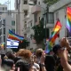 L'image montre une scène vibrante de célébration lors d'un défilé de fierté. On y voit une foule de personnes brandissant des drapeaux arc-en-ciel, symboles de la communauté LGBTQ+. Parmi eux, une grande bannière représentant le drapeau d'Israël flotte également. En arrière-plan, on distingue des bâtiments urbains, et plusieurs personnes semblent s'amuser et célébrer l'événement, créant une ambiance festive et joyeuse.