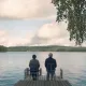 L'image montre deux personnes assises sur des chaises en bois sur une jetée qui s'avance dans un lac paisible. Ils regardent vers l'horizon, avec des arbres verdoyants en arrière-plan et un ciel nuageux au-dessus. L'atmosphère est calme et sereine, évoquant un moment de contemplation ou de discussion tranquille.