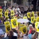 L'image montre une grande foule de personnes participant à une manifestation. Elles tiennent des affiches jaunes avec un symbole de main noire en faisant le signe qui pourrait représenter un mouvement ou une cause spécifique. La foule semble déterminée et engagée, et l'atmosphère est chargée d'énergie. On peut voir des pancartes et des slogans en arrière-plan. L'événement semble vouloir exprimer une opinion ou revendiquer des droits.