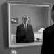 L'image montre un homme en costume noir se tenant devant un miroir. Son reflet dans le miroir affiche une expression sérieuse. L'environnement semble sobre, avec des éléments de décoration tels qu'une lampe et un cadre au mur. La scène est présentée en noir et blanc, ajoutant une atmosphère mélancolique et introspective à l'image.