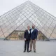 L'image montre deux hommes posant ensemble devant le Musée du Louvre, qui est célèbre pour sa pyramide en verre. Le bâtiment est moderne et impressionnant, contrastant avec l'architecture classique des ailes du musée. Les hommes sourient et portent des costumes formels, se tenant côte à côte, tandis que le ciel est légèrement nuageux, ajoutant une atmosphère calme à la scène.