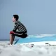 L'image montre un jeune garçon assis sur une coque de bateau échouée sur une plage. Il porte un t-shirt à rayures et un pantalon rouge. L'environnement est clair, avec un ciel bleu pâle et une plage de sable. Le garçon semble pensif, tourné vers l'horizon, tandis que la coque du bateau, abandonnée et usée, ajoute un élément de mélancolie à la scène.