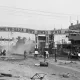 L'image montre une rue dans une ville, probablement à Gaza, avec une grande arche à l'entrée où il est écrit "Welcome to Gaza" en plusieurs langues. La scène est en noir et blanc, ce qui donne une atmosphère nostalgique ou dramatique. On aperçoit quelques personnes sur des bicyclettes et d'autres regroupées sur le côté de la route. Le décor urbain semble en désordre, avec des bâtiments qui montrent des signes d'usure et de dévastation. De la poussière s'élève, ajoutant à l'ambiance de la scène.