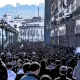 L'image montre une foule dense de personnes marchant dans une rue, probablement dans une ville animée. Les bâtiments sur les côtés semblent historiques, avec une architecture riche. On peut voir des lampadaires le long de la promenade, et au loin, on aperçoit des collines ou des montagnes qui ajoutent à la profondeur de la scène. L'atmosphère semble vibrante et dynamique, reflétant probablement une manifestation ou un événement public.