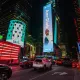 L'image montre une scène animée de Times Square à New York, illuminée par de nombreux panneaux publicitaires lumineux. Au centre de l'image, on peut voir une grande affiche mettant en avant une personne, Amr Diab, avec un sourire. Les bâtiments environnants sont recouverts de publicités colorées, dont l'une représente un drapeau américain. On aperçoit également des voitures circulant dans la rue, ajoutant à l'ambiance dynamique de ce célèbre endroit. Les lumières vives et les écrans créent une atmosphère vibrante et énergique.