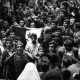 L'image montre une foule dense de personnes rassemblées dans une rue. Au premier plan, plusieurs individus sont visibles, dont un homme qui semble porter un drapeau avec un symbole distinctif. Les expressions des visages varient, certains levant les bras en signe de soutien ou d'enthousiasme. On peut observer des jeunes, des adultes et même des enfants parmi la foule. L'atmosphère semble intense et chargée d'émotion, indiquant un moment significatif ou une manifestation. La scène est en noir et blanc, ce qui lui donne un aspect historique.