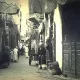 L'image montre une ruelle étroite d'une ville ancienne, probablement dans un pays arabe. Les murs des bâtiments sont ornés de tentures et de draps qui ajoutent une ambiance colorée. On peut voir des habitants, dont certains portent des vêtements traditionnels, déambulant dans la rue. Un homme semble se tenir près d'une charrette, tandis que d'autres personnes se déplacent en arrière-plan. Les portes en bois, typiques, ajoutent un charme ancien à la scène, qui évoque une atmosphère de vie quotidienne dans un vieux quartier.