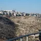 L'image montre un paysage caractérisé par une colline aride et rocheuse, sur laquelle se trouvent des bâtiments modernes. En arrière-plan, on peut voir des constructions en cours, avec des grues qui ajoutent à l'atmosphère de développement urbain. Au premier plan, une structure en béton est visible, semblant faire partie d'une voie ou d'un pont qui traverse le terrain. Le ciel est dégagé, ce qui donne une impression de luminosité et d'espace.