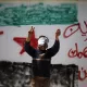 L'image montre un jeune garçon, les yeux bandés, levant les bras en signe de victoire ou de protestation. Il se trouve devant un mur sur lequel sont inscrits des slogans en arabe, ainsi qu'un symbole représentant le drapeau syrien. Cette scène évoque des thèmes de résistance et de luttes dans un contexte de conflit.