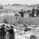 L'image montre une vue panoramique d'un paysage urbain historique. Au premier plan, un groupe de personnes se tient sur un balcon ou une terrasse, observant la scène devant eux. On aperçoit des ruines et des bâtiments anciens, tandis qu'en arrière-plan, on distingue des collines et des arbres. La célèbre mosquée du Dôme du Rocher est visible, ajoutant une dimension significative à cette scène. Le ciel est partiellement nuageux, et l'image évoque une atmosphère de calme et de contemplation.