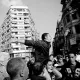 L'image montre une scène de manifestation dans un environnement urbain. Au centre, un homme se tient debout, les bras écartés, semblant exprimer une idée forte ou appeler à l'action. Il est entouré d'une foule dense, composée d'hommes, qui l'écoute ou réagit à son discours. Les bâtiments en arrière-plan montrent une architecture typique de ville, et l'image est en noir et blanc, ce qui accentue les émotions et la tension de la situation. L'atmosphère semble dynamique et engageante.