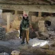 L'image montre un intérieur d'un bâtiment qui semble ancien et en ruine. On peut voir des murs en pierre et des débris éparpillés sur le sol. Un homme, habillé en vêtement militaire avec un bandeau, se tient au centre de la pièce, tandis que deux autres personnes, cachées en partie, l'observent. La lumière entre par des ouvertures, créant un contraste entre les ombres et les zones illuminées dans cet environnement désolé.