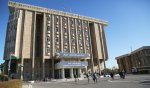 L'image montre un bâtiment imposant, probablement un édifice institutionnel, avec des drapeaux flottant à l'extérieur. La façade du bâtiment est en pierre avec un design moderne, et il est clairement identifié par une inscription en arabe et en anglais indiquant qu'il s'agit du "Parlement du Kurdistan - Irak". Il y a des personnes qui se déplacent autour du bâtiment, ce qui évoque un lieu d'activité politique et administrative. L'environnement semble clair et ensoleillé, avec un ciel bleu en arrière-plan.