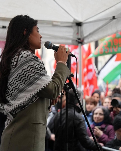 L'image montre une femme se tenant devant un microphone, probablement en train de prendre la parole lors d'une manifestation. Elle porte un manteau et un foulard avec un motif traditionnel. En arrière-plan, on aperçoit une foule portant des drapeaux et des pancartes, ce qui indique que l'événement est lié à une cause politique ou sociale. L'atmosphère semble engagée et mobilisée.