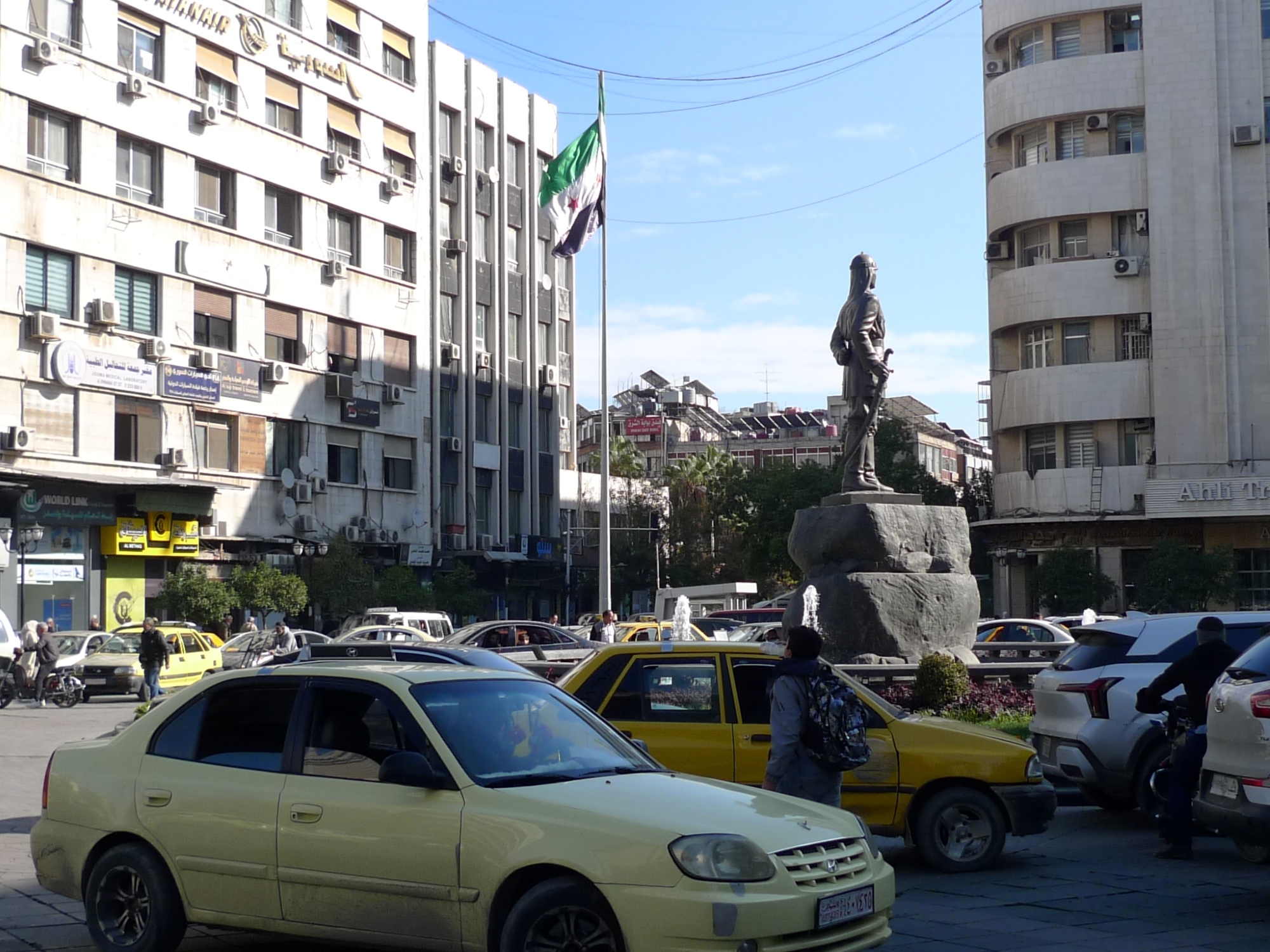 Une place urbaine avec des voitures et une statue près d'un drapeau.