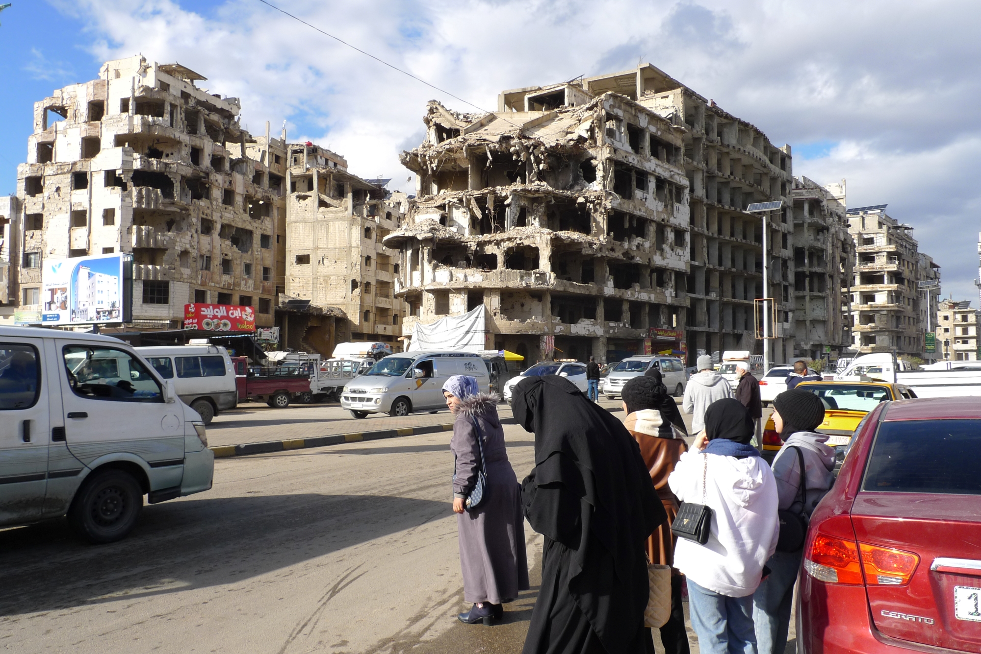 Des bâtiments détruits, en ruines, avec des personnes attendant au bord de la route.