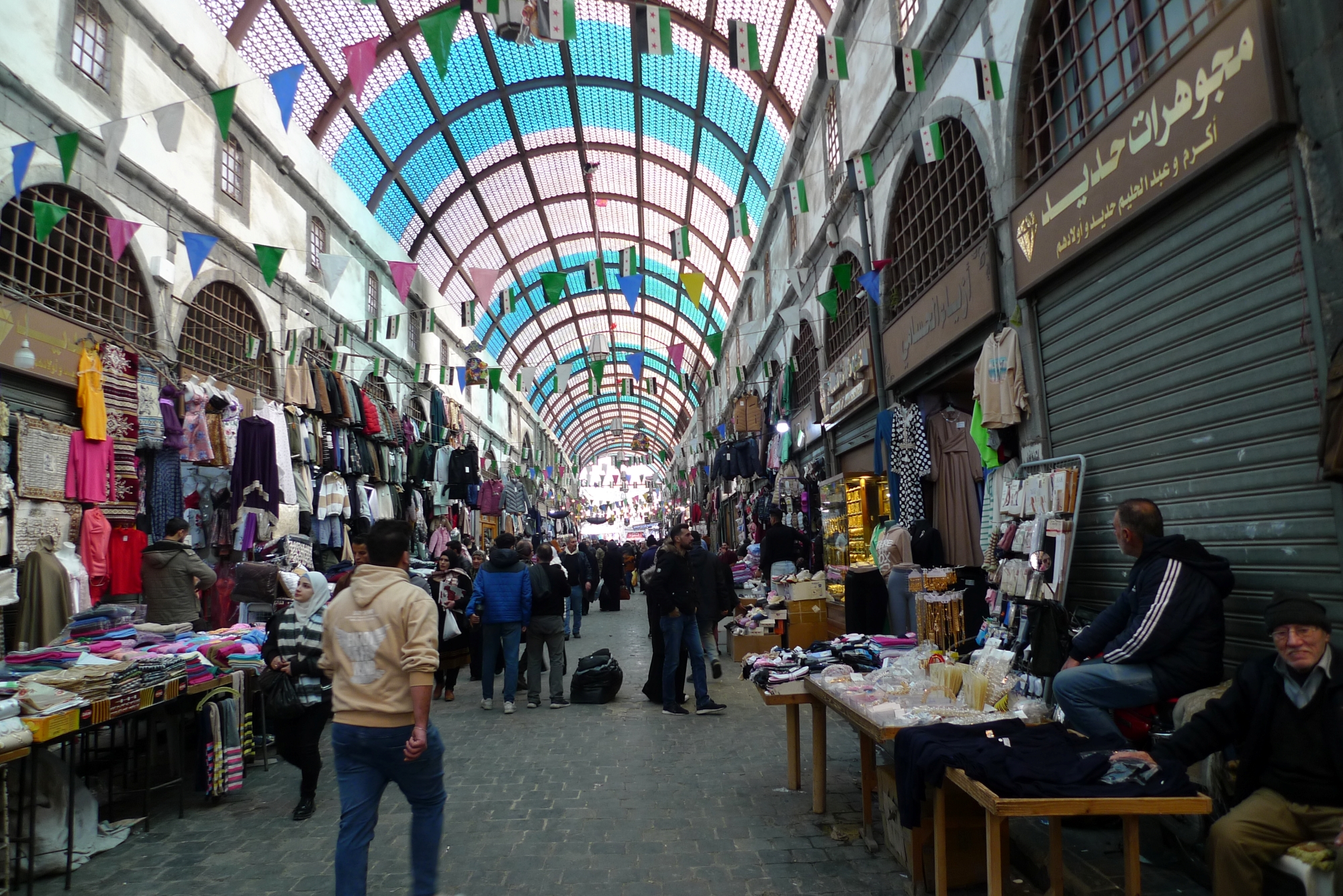 Un marché animé, avec des vêtements, des étals et des gens qui se déplacent sous un toit coloré.