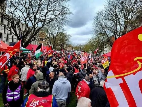 L'image montre une manifestation animée. On peut y voir une foule rassemblée, avec de nombreuses personnes brandissant des drapeaux de différentes couleurs et emblèmes, en particulier ceux de la CGT (Confédération Générale du Travail). L'ambiance semble dynamique et mobilisée, avec des arbres dénudés en arrière-plan et un ciel nuageux. Les participants portent des vêtements variés, et l'atmosphère suggère une lutte ou une revendication collective.
