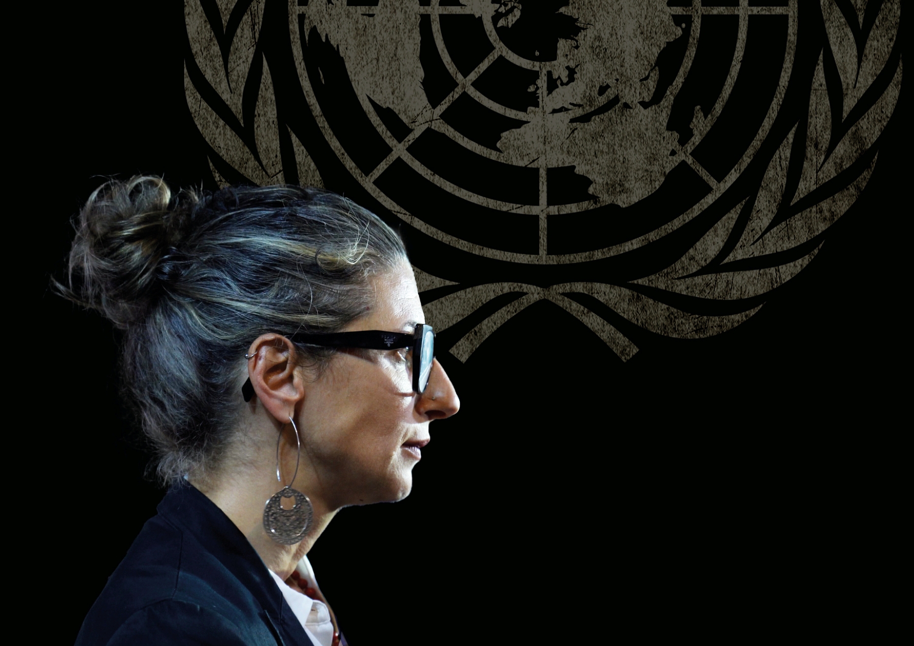 زنی با موهای خاکستری و عینک، در کنار نماد سازمان ملل، در حال صحبت کردن است.