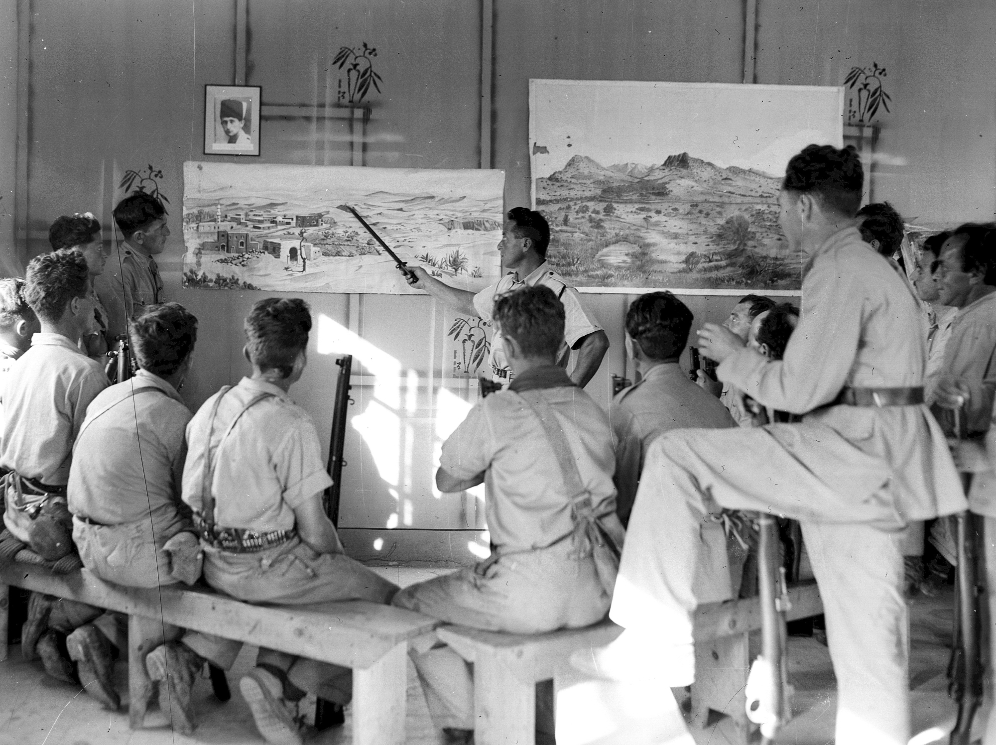 L'image montre un groupe de personnes assises sur des bancs, attentives à un homme qui semble donner une présentation devant des tableaux accrochés au mur. Les tableaux illustrent des paysages, l'un représentant probablement un village ou une scène de la nature. L'environnement est dépouillé, avec des murs simples et une lumière naturelle qui pénètre à travers une fenêtre. Les participants, majoritairement en vêtements uniformes, semblent captivés par l'explication de l'intervenant. Il y a aussi un portrait encadré sur le mur, ajoutant un élément de contexte à la scène.