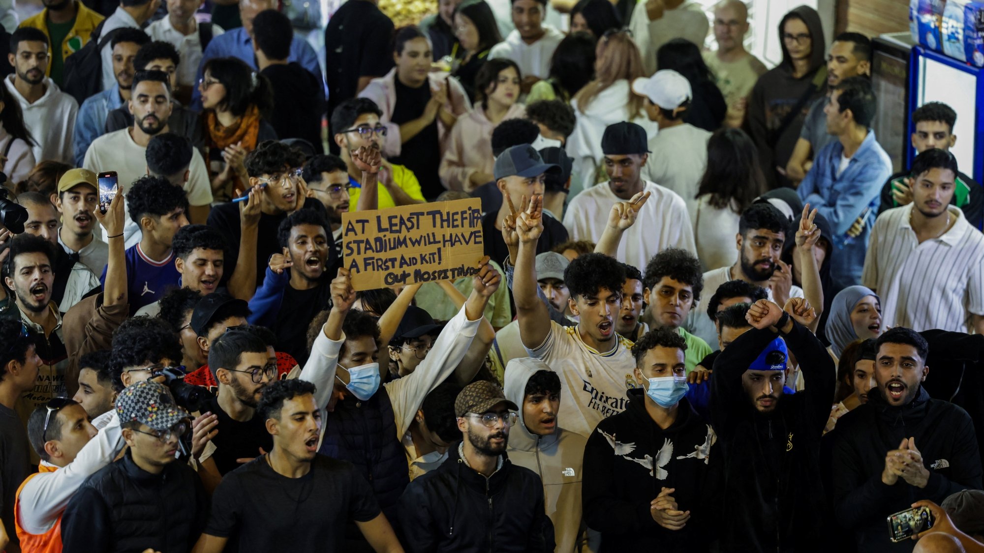 حشود من الناس في مكان مزدحم، يرفعون لافتة ويهتفون، مع تباين في الألوان والملابس.