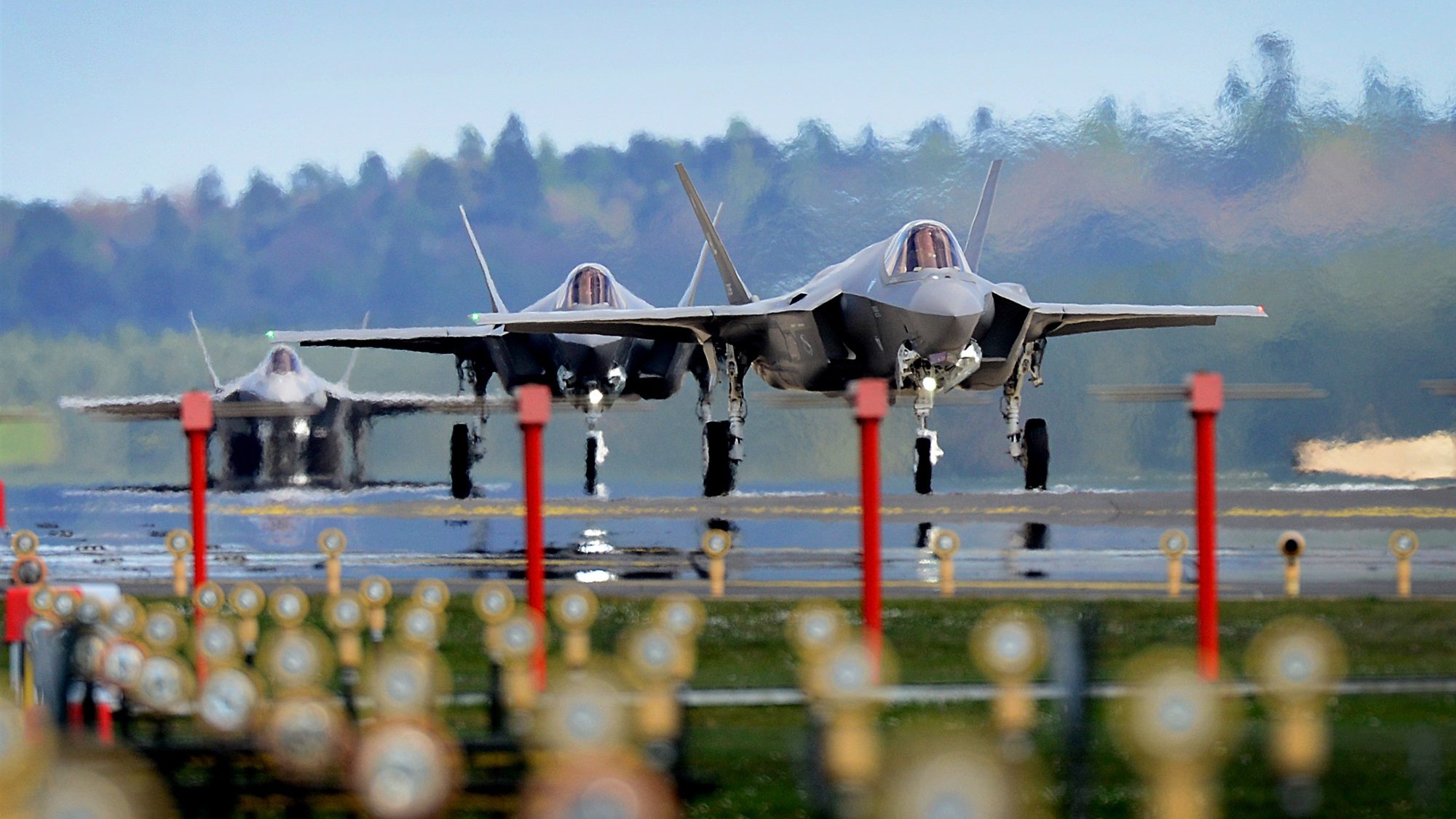 تظهر في الصورة ثلاث طائرات مقاتلة من طراز F-35 تتواجد على مدرج مطار. الطائرات تبدو وكأنها جاهزة للإقلاع، حيث تبرز من الخلف ملامحها الحديثة وتصميمها الأنيق. الأرضية محاطة بمجموعة من الحواجز أو الأعمدة، في حين أن المناظر الطبيعية في الخلفية تظهر أشجارًا ومساحات خضراء تحت سماء زرقاء. تُعطي الصورة انطباعًا عن القوة والتقنية المتقدمة في مجال الطيران العسكري.