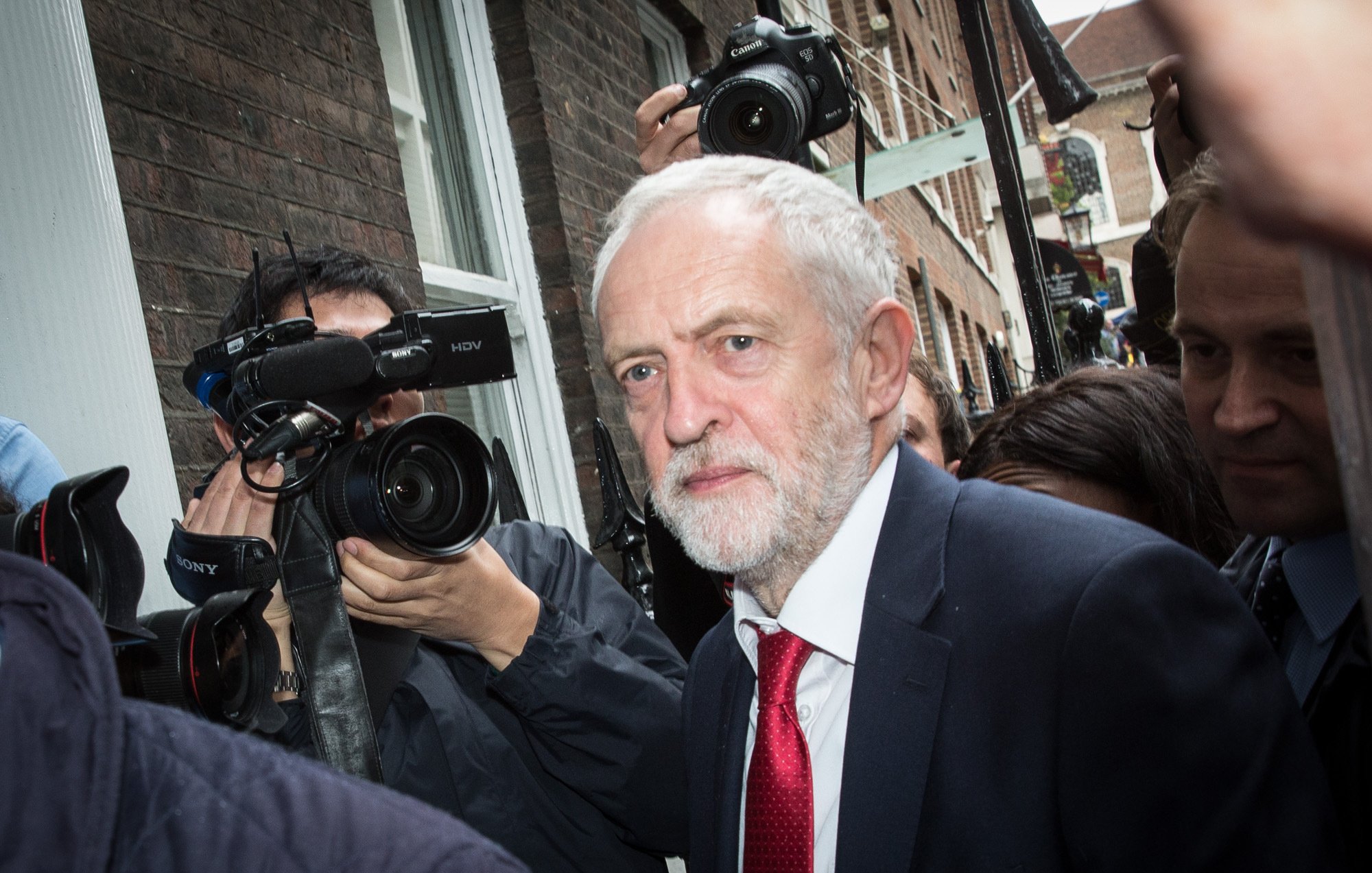 الصورة تظهر رجلًا متوسط العمر ذو لحية وشعر رمادي، يرتدي بدلة داكنة وربطة عنق حمراء. يبدو أنه في موقف يتطلب الانتباه، حيث تحيط به مجموعة من الكاميرات والصحفيين الذين يلتقطون صورًا له. الخلفية تعكس بيئة حضرية مع مبانٍ طوبية، وتعكس الحركة والاهتمام بالشخص.