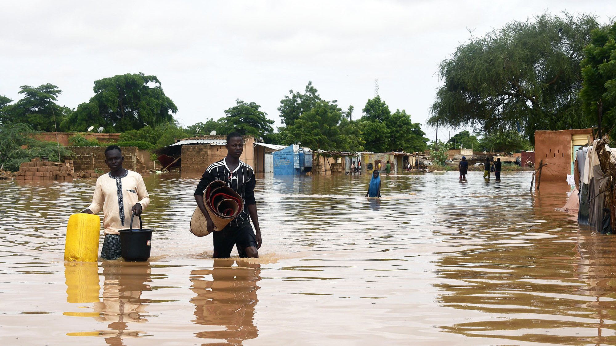تظهر الصورة مشهداً لمنطقة تعرضت للفيضانات، حيث يغمر الماء الأرض بشكل كبير. يوجد شخصان يسيران في المياه العميقة، يحمل أحدهما دلوًا ويبدو أنهما يجمعان بعض الممتلكات. في الخلفية، يمكن رؤية أكواخ وأشجار، مما يدل على أن المنطقة قد تضررت. السماء ملبدة بالغيوم، مما يشير إلى ظروف جوية غير مستقرة. يعكس المشهد الصعوبات التي يواجهها السكان نتيجة الفيضانات.